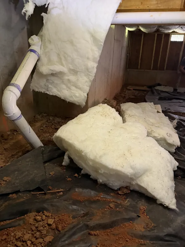 Chunks of fallen fiberglass insulation on torn black plastic and red clay beside a white drain pipe